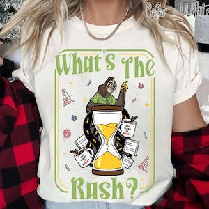 Puede incluir: Camiseta blanca con un gr&aacute;fico humor&iacute;stico que presenta un perezoso, un reloj de arena y el texto "What's The Rush?". El dise&ntilde;o incluye tazas de caf&eacute; y elementos de carretera, creando un tema l&uacute;dico. La camiseta es de material suave.