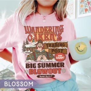 Op de afbeelding: Lichtroze T-shirt met een retro graphic. Het ontwerp luidt "Wandering Oaken's Trading Post and Sauna! Big Summer Blowout" met een cartoonfiguur en een hut. Een mok met een smiley wordt in de afbeelding vastgehouden.