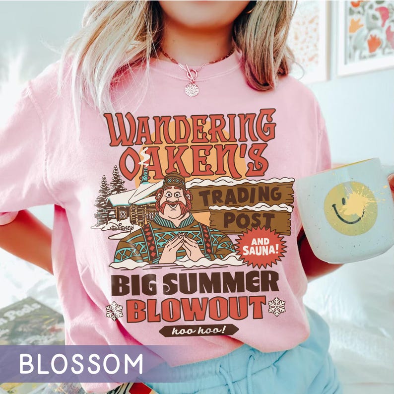 Op de afbeelding: Lichtroze T-shirt met een retro graphic. Het ontwerp luidt "Wandering Oaken's Trading Post and Sauna! Big Summer Blowout" met een cartoonfiguur en een hut. Een mok met een smiley wordt in de afbeelding vastgehouden.