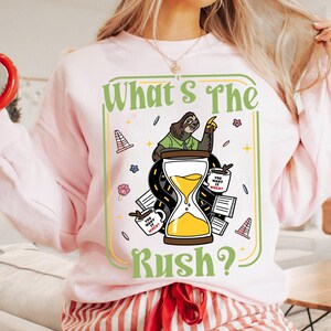 Puede incluir: Sudadera rosa claro con un dise&ntilde;o gr&aacute;fico humor&iacute;stico. Presenta un perezoso, un reloj de arena y la frase "What's The Rush?". El dise&ntilde;o incluye tazas de caf&eacute; y elementos de carretera.