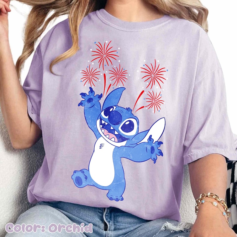 May include: Orchid-colored t-shirt featuring a blue cartoon character with fireworks. The character is smiling with its arms raised, and the fireworks are red and white. The text "Color: Orchid" is at the bottom.