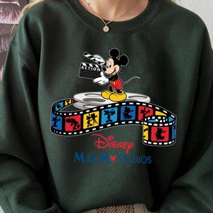 Pu&ograve; includere: Felpa verde scuro con Topolino che tiene una clapperboard con la scritta "ACTION". Il design include una bobina di film e una striscia di film colorata con il testo Disney MGM Studios.