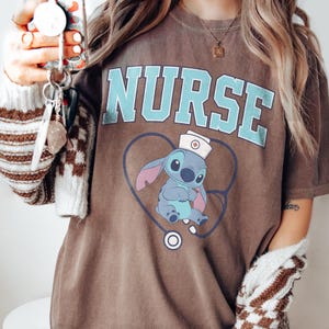 May include: Brown t-shirt with the word "NURSE" in light blue block letters. Below the word is a cartoon character wearing a nurse's hat and stethoscope. The shirt is a casual style.