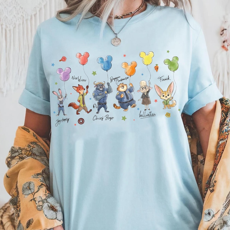 Op de afbeelding: Lichtblauw T-shirt met Zootopia-personages die Mickey Mouse-vormige ballonnen vasthouden. Het ontwerp bevat Judy Hopps, Nick Wilde, Chief Bogo en anderen. De namen van de personages staan eronder.