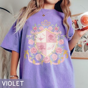 Puede incluir: Camiseta violeta con un dise&ntilde;o de escudo con una corona, rosas y cintas. El escudo incluye una l&aacute;mpara, una concha, una flor de loto y un zapato de cristal. La palabra "VIOLET" est&aacute; impresa en la parte inferior.