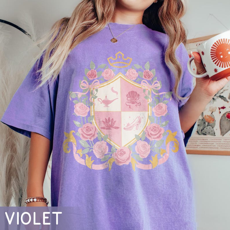 Puede incluir: Camiseta violeta con un dise&ntilde;o de escudo con una corona, rosas y cintas. El escudo incluye una l&aacute;mpara, una concha, una flor de loto y un zapato de cristal. La palabra "VIOLET" est&aacute; impresa en la parte inferior.