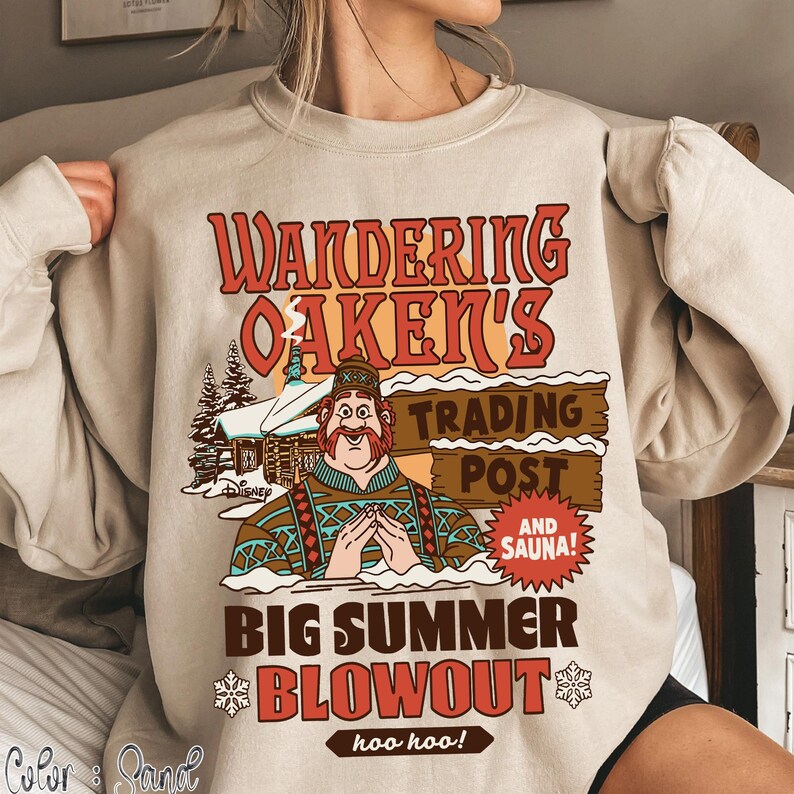Op de afbeelding: Beige sweatshirt met een afbeelding met de tekst "Wandering Oaken's Trading Post and Sauna!" in rood en bruin. De afbeelding bevat een cartoon van een man en een besneeuwde hut. De zin "Big Summer Blowout" wordt ook weergegeven.
