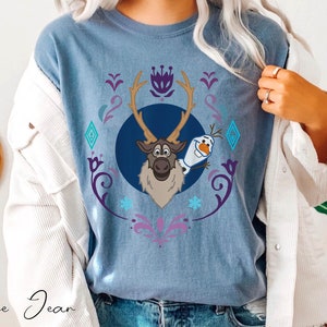 Puede incluir: Camiseta azul vaquero con un gr&aacute;fico de Sven el reno y Olaf el mu&ntilde;eco de nieve de la pel&iacute;cula Frozen. El dise&ntilde;o est&aacute; enmarcado por un patr&oacute;n floral y geom&eacute;trico decorativo en morado, azul y blanco. Las palabras "Blue Jean" est&aacute;n escritas en cursiva en la parte inferior.
