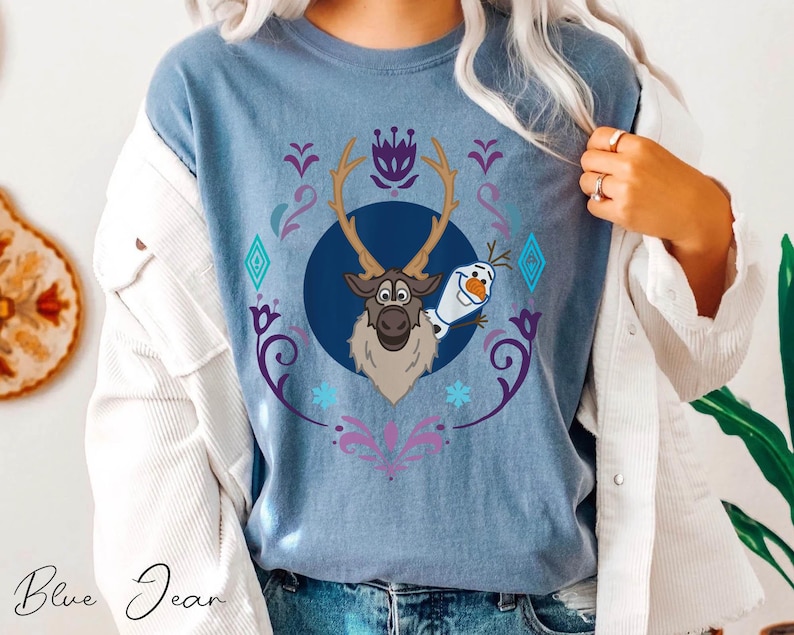 Puede incluir: Camiseta azul vaquero con un gr&aacute;fico de Sven el reno y Olaf el mu&ntilde;eco de nieve de la pel&iacute;cula Frozen. El dise&ntilde;o est&aacute; enmarcado por un patr&oacute;n floral y geom&eacute;trico decorativo en morado, azul y blanco. Las palabras "Blue Jean" est&aacute;n escritas en cursiva en la parte inferior.