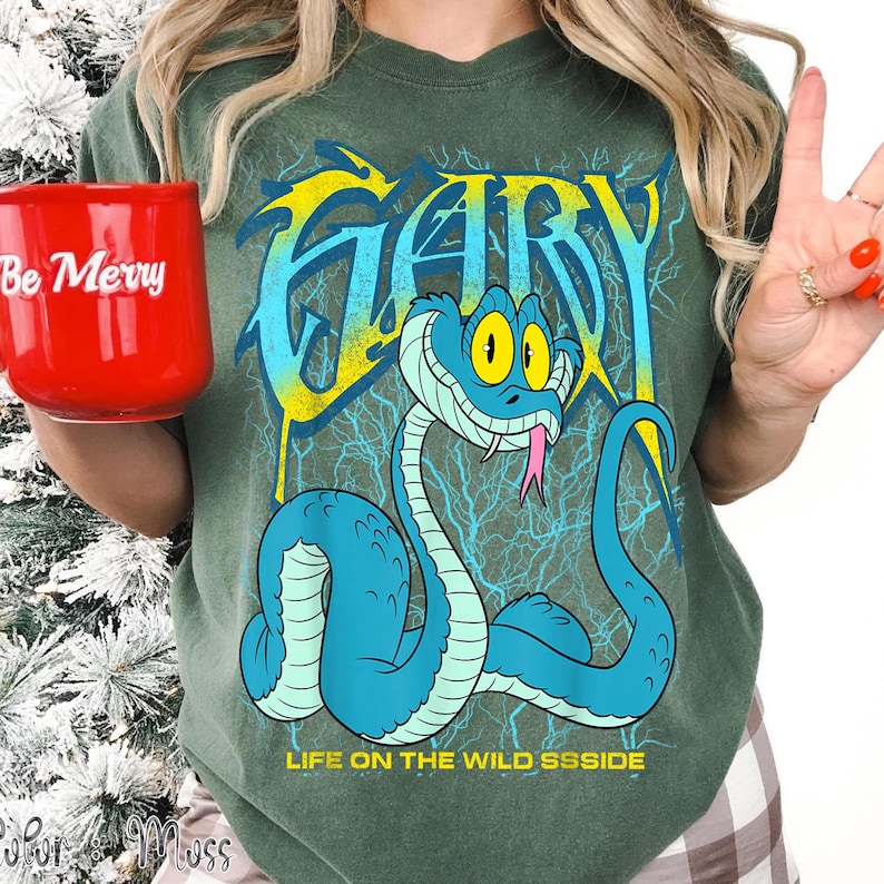 Op de afbeelding: Een groen t-shirt met een cartoon slang ontwerp. Het woord "GARY" staat in geel en blauw, met een blauwe slang eronder. De slang heeft een roze tong. De tekst "LIFE ON THE WILD SSSSIDE" staat eronder. Een rode mok met "Be Merry" wordt vastgehouden.