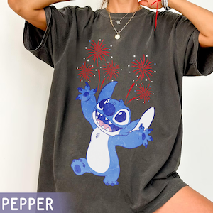 May include: Dark gray t-shirt featuring a blue cartoon character with fireworks. The character is blue with white accents and a large grin. Red and white fireworks burst above the character. The word "PEPPER" is at the bottom.
