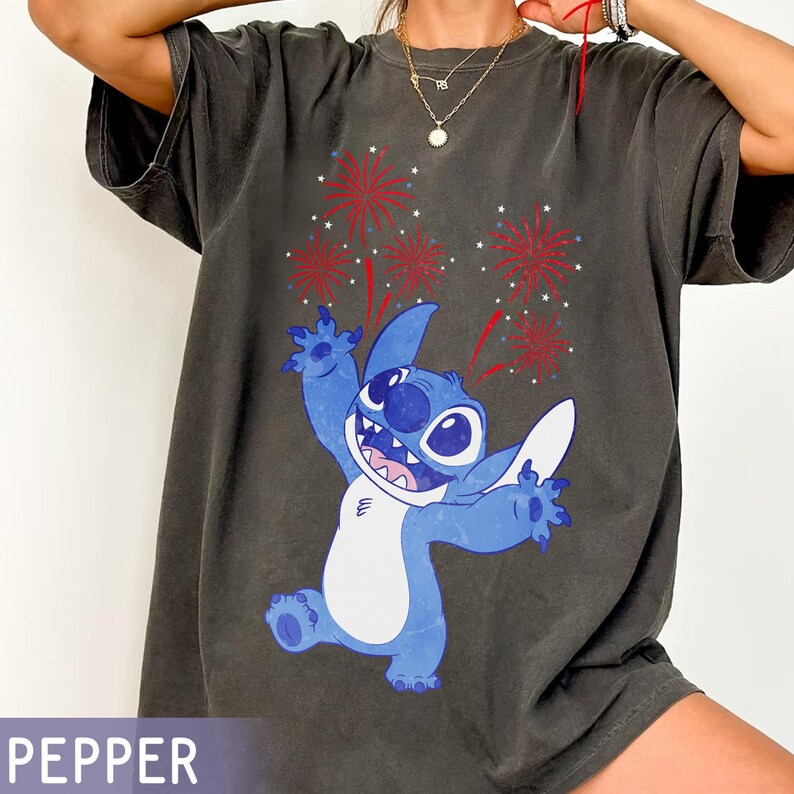 May include: Dark gray t-shirt featuring a blue cartoon character with fireworks. The character is blue with white accents and a large grin. Red and white fireworks burst above the character. The word "PEPPER" is at the bottom.