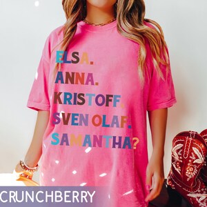 以下が含まれることがあります： エルサ、アナ、クリストフ、スヴェン、オラフ、サマンサ？という名前が様々な色でプリントされたピンク色のTシャツ。Tシャツはクルーネックで半袖です。画像の下部には「CRUNCHBERRY」という文字があります。