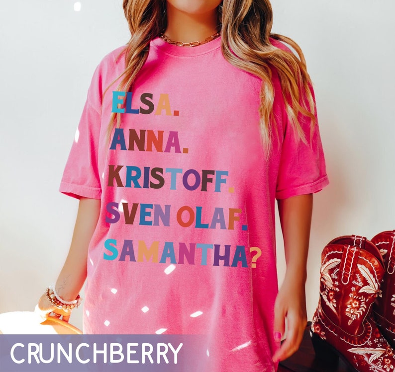 以下が含まれることがあります： エルサ、アナ、クリストフ、スヴェン、オラフ、サマンサ？という名前が様々な色でプリントされたピンク色のTシャツ。Tシャツはクルーネックで半袖です。画像の下部には「CRUNCHBERRY」という文字があります。