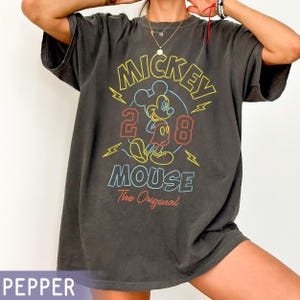 Puede incluir: Camiseta gris oscuro con un gr&aacute;fico colorido de Mickey Mouse y el texto "Mickey Mouse 2008 The Original". El dise&ntilde;o incluye detalles en amarillo, rojo y azul. La camiseta tiene un aspecto vintage y desgastado.