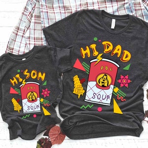 May include: Two grey t-shirts featuring a cartoon dog and a can of soup. The larger shirt reads "Hi Dad" and the smaller shirt reads "Hi Son". Both shirts have a retro 90s design with colourful geometric shapes.
