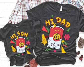 May include: Two grey t-shirts featuring a cartoon dog and a can of soup. The larger shirt reads "Hi Dad" and the smaller shirt reads "Hi Son". Both shirts have a retro 90s design with colourful geometric shapes.