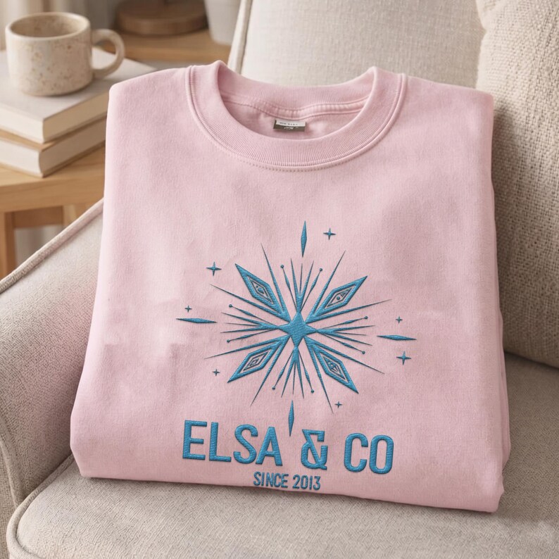 May include: A light pink crewneck sweatshirt featuring an embroidered blue snowflake design centered on the chest. Below the snowflake, the text reads ELSA & CO in blue capital letters, with the smaller text SINCE 2013 printed underneath.