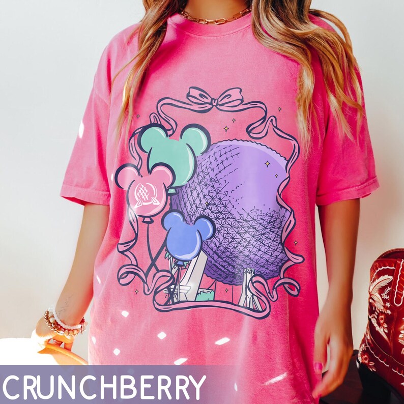 May include: A pink t-shirt with a graphic featuring a purple sphere, Mickey Mouse-shaped balloons in blue and green, and a ribbon design. The text "CRUNCHBERRY" is at the bottom of the shirt. The shirt is a short-sleeved, casual style.