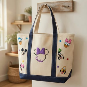 May include: A cream-colored canvas tote bag with navy blue handles and a navy blue base. The bag features embroidered patches of Disney characters including Mickey Mouse, Minnie Mouse, Donald Duck, Daisy Duck, Goofy, and Pluto, plus a central bow.