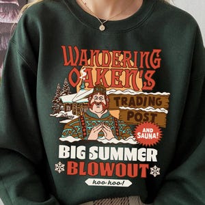 Op de afbeelding: Donkergroene sweatshirt met een afbeelding met de tekst "Wandering Oaken's Trading Post and Sauna!" en een cartoonafbeelding van een man. De tekst "Big Summer Blowout" wordt ook weergegeven, samen met sneeuwvlokken en de zin "hoo hoo!"