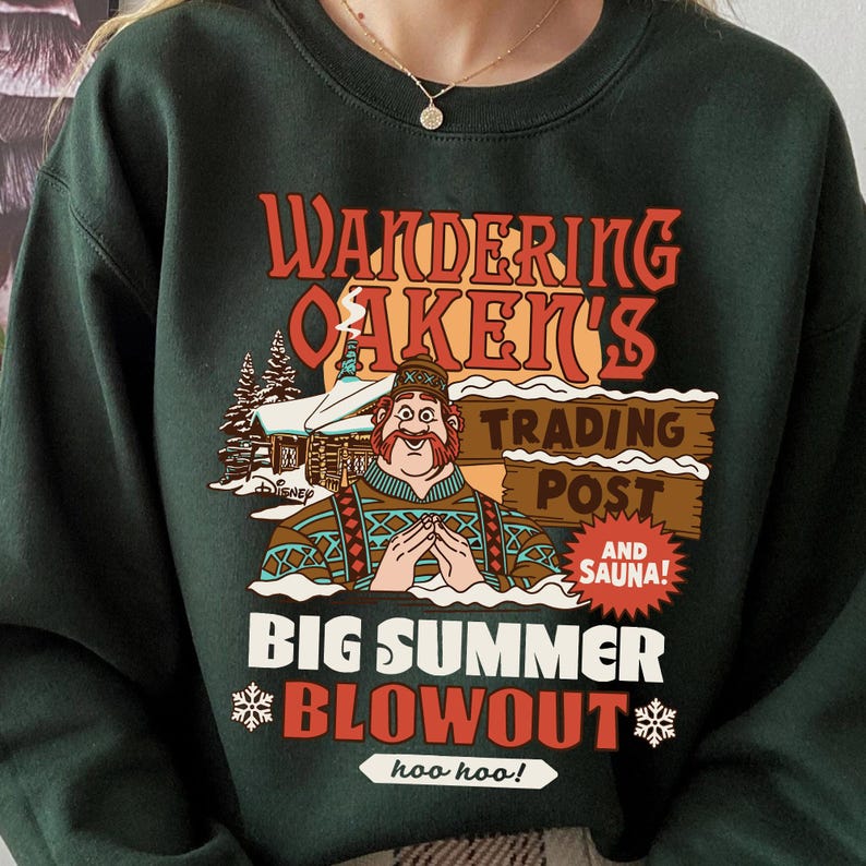 Op de afbeelding: Donkergroene sweatshirt met een afbeelding met de tekst "Wandering Oaken's Trading Post and Sauna!" en een cartoonafbeelding van een man. De tekst "Big Summer Blowout" wordt ook weergegeven, samen met sneeuwvlokken en de zin "hoo hoo!"