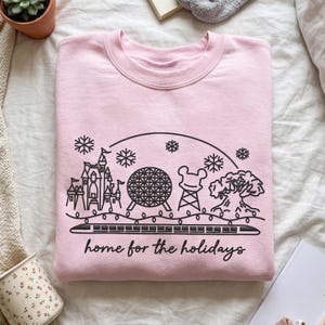 May include: A light pink crewneck sweatshirt featuring a black line art graphic of a theme park skyline with a castle, a geodesic sphere, a tower, and a tree. The design includes snowflakes, string lights, a monorail, and the text home for the holidays.