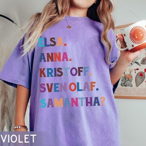 以下が含まれることがあります： エルサ、アナ、クリストフ、スヴェン、オラフ、サマンサの名前が様々な色でプリントされた紫色のTシャツ。「VIOLET」の文字がシャツの下部にプリントされています。