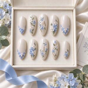 May include: A set of ten almond-shaped press-on nails in a cream base color, decorated with hand-painted blue and white floral patterns, small white pearls, and gold-colored letters A and J, all arranged neatly inside a square cream-colored box.