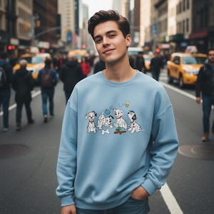 May include: A light blue sweatshirt featuring a graphic of five Dalmatian puppies playing with a ball and a bone, with a small bee. The sweatshirt is worn by a person standing on a city street.