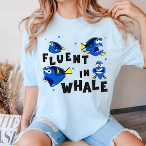 May include: A light blue t-shirt with a graphic of Dory from Finding Nemo. The shirt says "Fluent in Whale" with four images of Dory.
