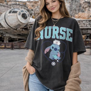May include: Dark gray t-shirt with the word "NURSE" in light blue block letters. Below the text is a cartoon image of Stitch wearing a nurse's hat inside a blue heart with a stethoscope. The shirt is a casual, oversized fit.