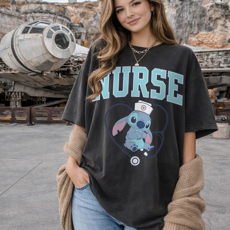 May include: Dark gray t-shirt with the word "NURSE" in light blue block letters. Below the text is a cartoon image of Stitch wearing a nurse's hat inside a blue heart with a stethoscope. The shirt is a casual, oversized fit.