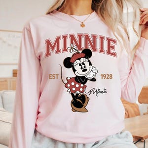 May include: A light pink long-sleeve sweatshirt featuring a Minnie Mouse graphic. The graphic includes the word "MINNIE" in a maroon collegiate font, the text "EST 1928", and a cartoon image of Minnie Mouse in a red polka dot dress.