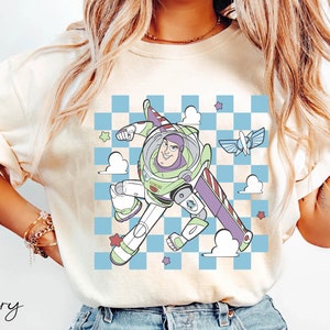 May include: A white t-shirt with a cartoon image of Buzz Lightyear from Toy Story flying in a checkered blue and white pattern with clouds and stars. The text "Ivory" is written in cursive at the bottom left corner of the image.