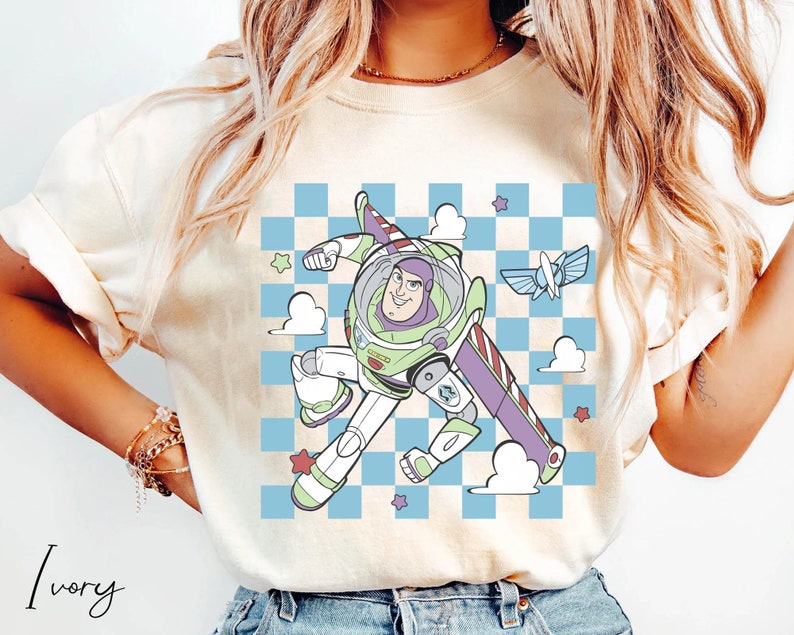 May include: A white t-shirt with a cartoon image of Buzz Lightyear from Toy Story flying in a checkered blue and white pattern with clouds and stars. The text "Ivory" is written in cursive at the bottom left corner of the image.
