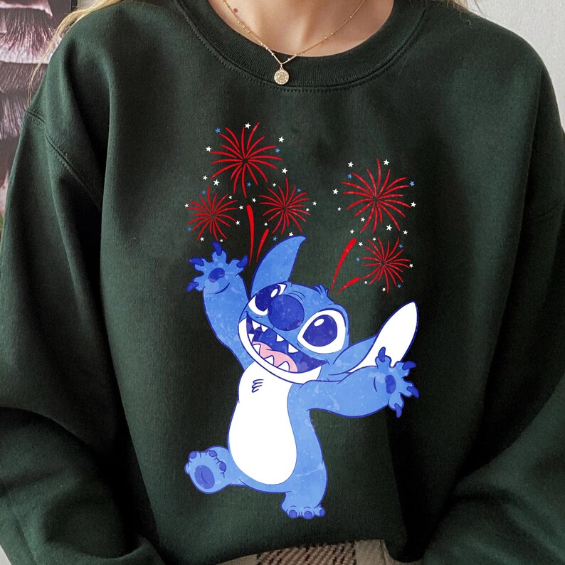 May include: Dark green sweatshirt featuring a blue cartoon character with large eyes and a white belly, celebrating with red and white fireworks. The character is smiling with its arms raised. A gold necklace is visible.