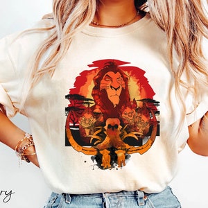 May include: A white t-shirt with a graphic print of Simba, the lion king, from the Disney movie The Lion King. The graphic features Simba with a red sunset background and a skull and crossbones in the foreground. The text "Ivory" is visible in the bottom left corner of the image.