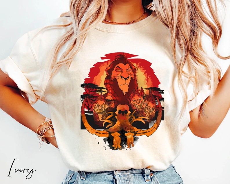 May include: A white t-shirt with a graphic print of Simba, the lion king, from the Disney movie The Lion King. The graphic features Simba with a red sunset background and a skull and crossbones in the foreground. The text "Ivory" is visible in the bottom left corner of the image.