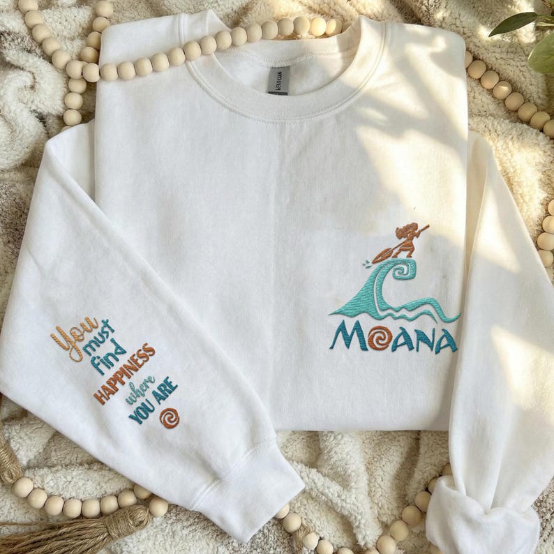 May include: A white crewneck sweatshirt featuring an embroidered design on the chest of a character paddling on a wave above the text MOANA. The sleeve features the embroidered text You must find HAPPINESS where YOU ARE with a spiral icon.
