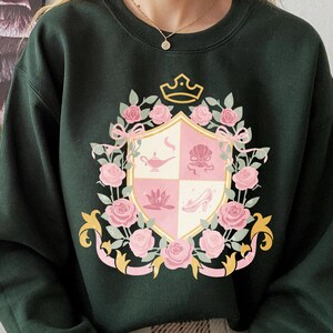 Puede incluir: Sudadera verde oscuro con un dise&ntilde;o de escudo de armas. El escudo presenta rosas rosas, lazos y una corona. El escudo est&aacute; dividido en cuatro secciones con elementos rosas y blancos, incluyendo una l&aacute;mpara, una concha, una flor de loto y un zapato de tac&oacute;n.