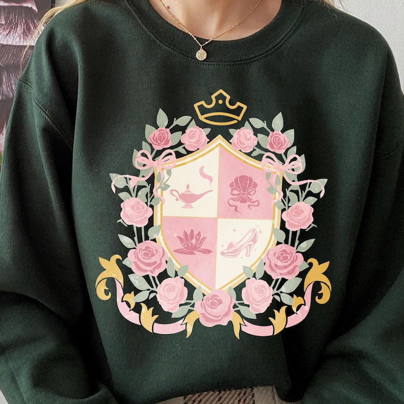Puede incluir: Sudadera verde oscuro con un dise&ntilde;o de escudo de armas. El escudo presenta rosas rosas, lazos y una corona. El escudo est&aacute; dividido en cuatro secciones con elementos rosas y blancos, incluyendo una l&aacute;mpara, una concha, una flor de loto y un zapato de tac&oacute;n.