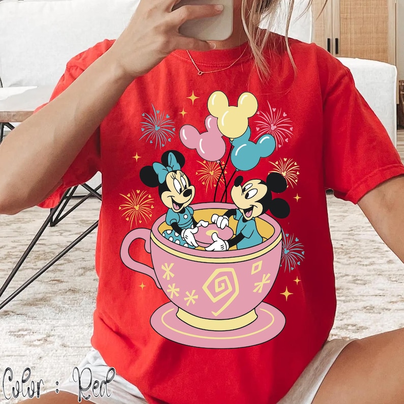 May include: Red t-shirt with a graphic of Mickey and Minnie Mouse in a teacup, holding balloons. The design includes fireworks and the text "Color: Red". The teacup is pink with yellow accents.