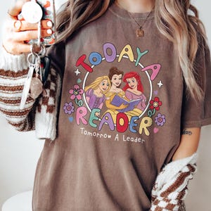 May include: A brown t-shirt with a colorful graphic featuring three cartoon princesses inside a circle. The text reads "Today a Reader, Tomorrow a Leader". The design includes flowers and stars.