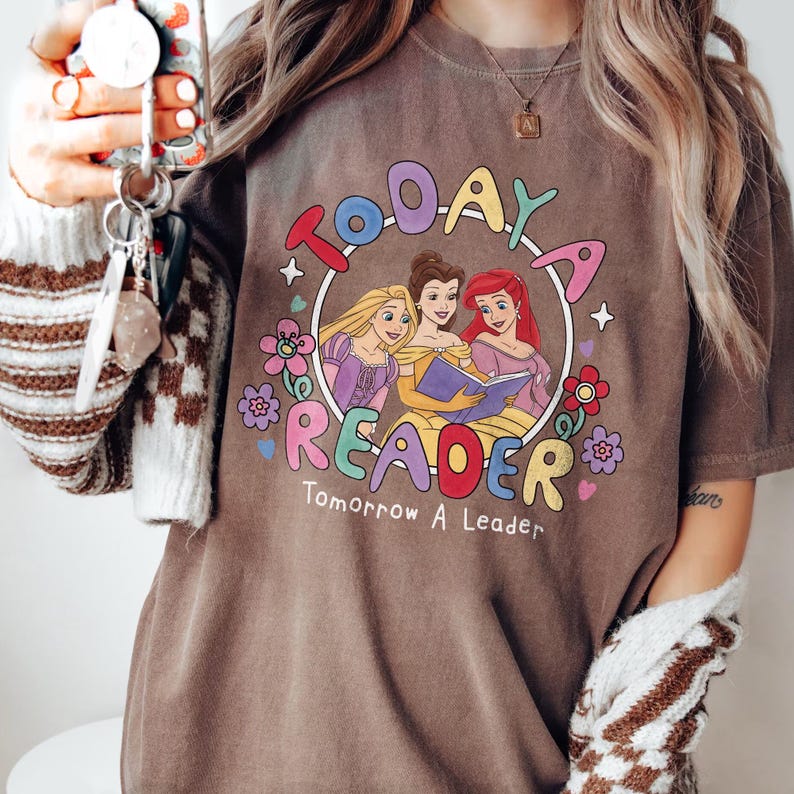 May include: A brown t-shirt with a colorful graphic featuring three cartoon princesses inside a circle. The text reads "Today a Reader, Tomorrow a Leader". The design includes flowers and stars.