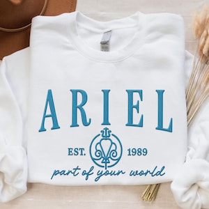 May include: White sweatshirt with the name "ARIEL" embroidered in teal, along with the text "EST. 1989" and "part of your world". A teal emblem is also featured. The sweatshirt is laid flat on a neutral surface.