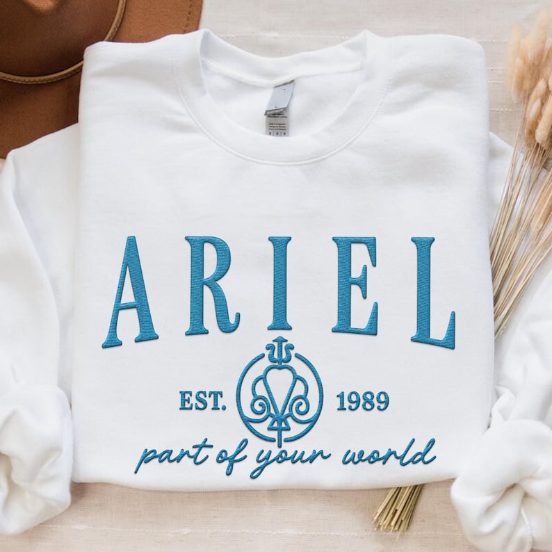 May include: White sweatshirt with the name "ARIEL" embroidered in teal, along with the text "EST. 1989" and "part of your world". A teal emblem is also featured. The sweatshirt is laid flat on a neutral surface.