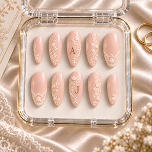 May include: A set of ten almond-shaped press-on nails in a soft blush pink hue, featuring intricate white 3D floral lace relief patterns. Two nails are accented with gold-colored letters A and J, while others display delicate lace borders.