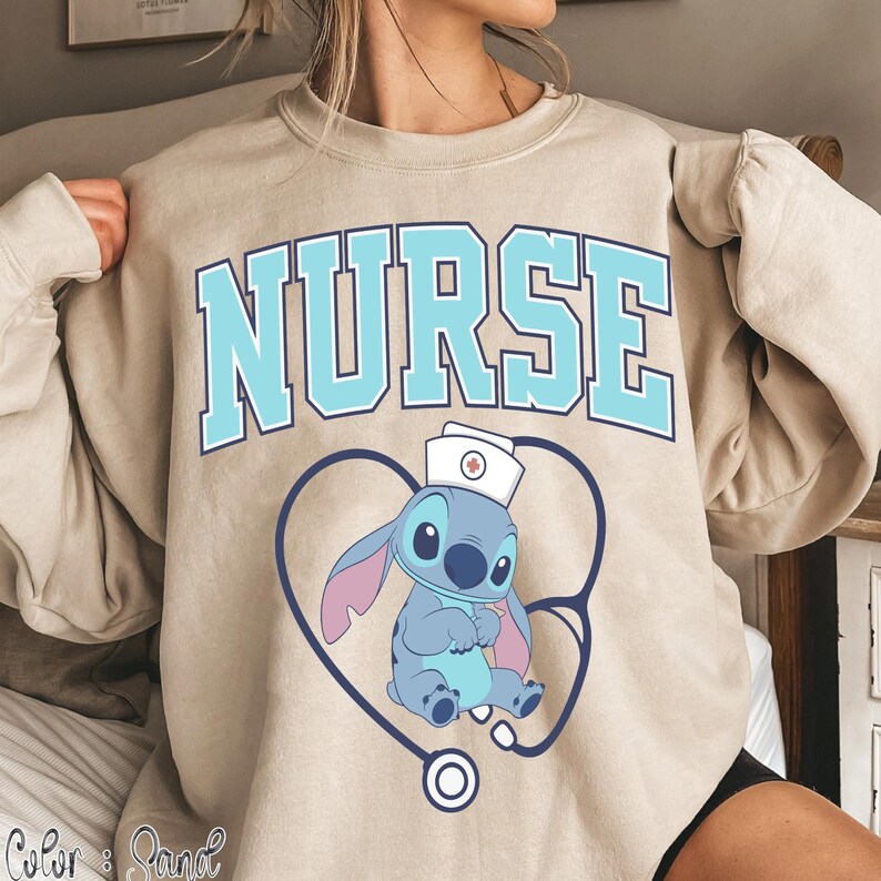 May include: Beige sweatshirt with the word "NURSE" in light blue block letters. A cartoon character wearing a nurse's hat is inside a blue stethoscope heart. The character is blue with pink ears.