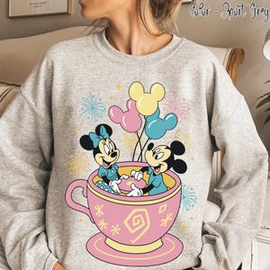 May include: Sport grey sweatshirt with a graphic of Mickey and Minnie Mouse in a pink teacup, holding a plate. The design includes balloons and fireworks, with the text "Color: Sport Grey".